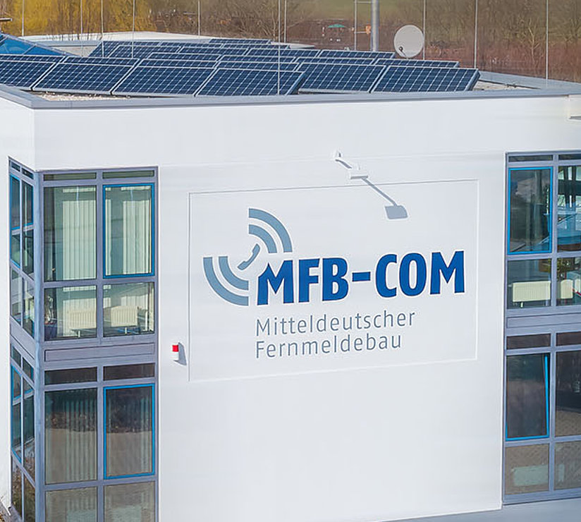 Ein modernes weißes Bürogebäude mit großen Fenstern und Sonnenkollektoren auf dem Dach, auf dem das MFB-COM-Logo prangt. Vor dem Gebäude sind mehrere weiße Lieferwagen geparkt. Der Text lautet Telekommunikation Neu Gedacht.