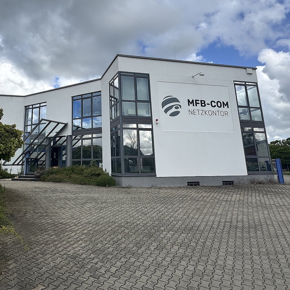 Ein modernes weißes Bürogebäude mit großen Fenstern und dem Schriftzug MFB-COM NETZKONTOR an der Fassade, vor einem wolkenverhangenen Himmel. In der Nähe des Eingangs gibt es Grünflächen und ein Schild mit Informationen zum Unternehmen.