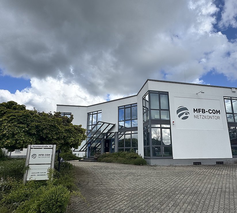 Ein modernes weißes Bürogebäude mit großen Fenstern und dem Schriftzug MFB-COM NETZKONTOR an der Fassade, vor einem wolkenverhangenen Himmel. In der Nähe des Eingangs gibt es Grünflächen und ein Schild mit Informationen zum Unternehmen.