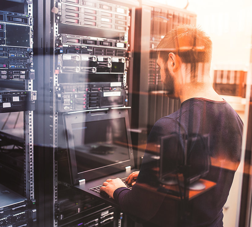 Ein Mann steht in einem Serverraum und arbeitet an einem Laptop, der an ein Server-Rack voller Geräte und Kabel angeschlossen ist. Durch eine Glaswand wird die Szene von einem warmen Licht erhellt.
